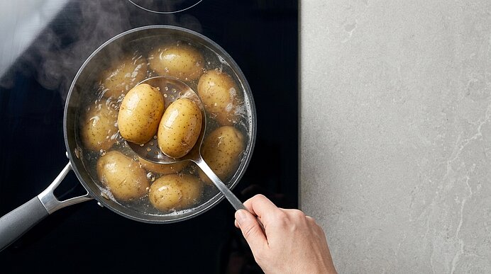 Hand mit Schaumlöffel hebt kleine Kartoffeln aus kochendem Wasser in einem Topf.