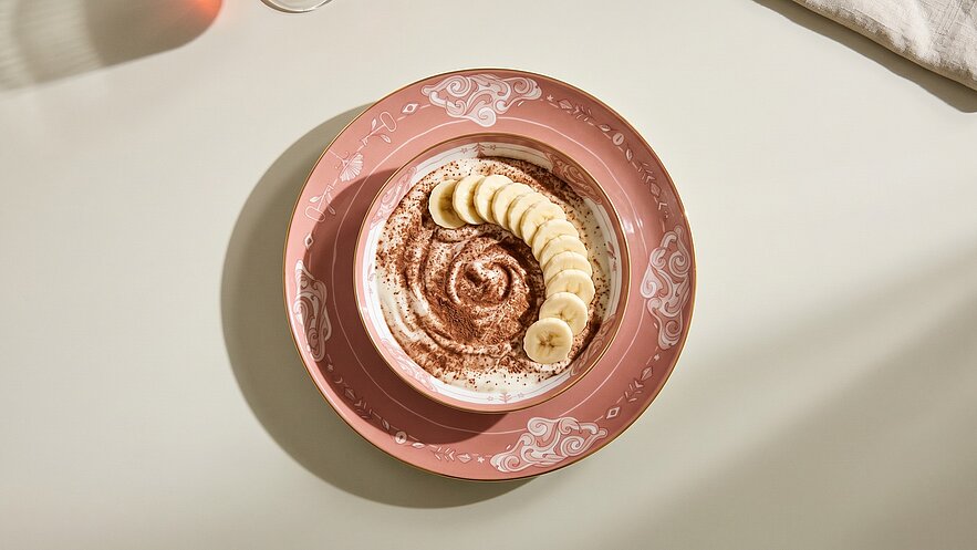 Eine rosa Schüssel mit einer cremigen Schicht, dekoriert mit Kakaopulver und Banane in Scheiben, die spiralartig angeordnet sind. Die Präsentation des Desserts gibt ihm ein ansprechendes Aussehen.