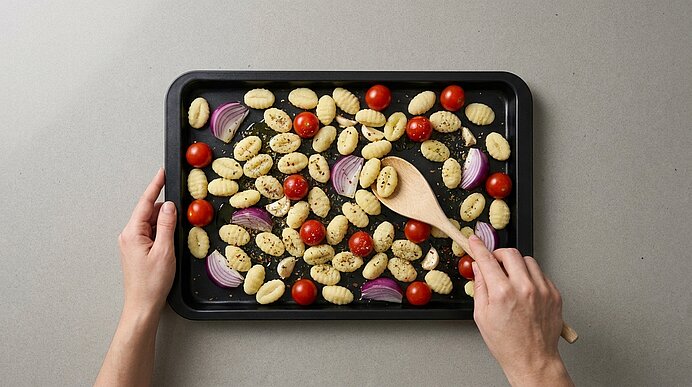 Backblech mit gewürzten Gnocchi, Kirschtomaten und roten Zwiebelstücken; eine Person rührt die Zutaten mit einem Holzlöffel.