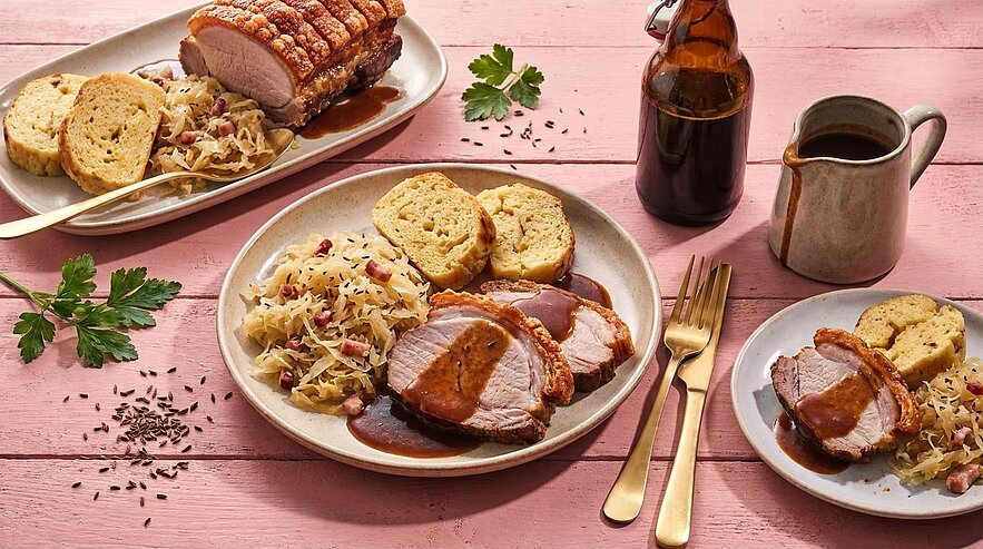 Teller mit in Scheiben geschnittenem Schweinebraten mit knuspriger Kruste, dunkler Bratensoße, Sauerkraut und Semmelknödeln; im Hintergrund Bräter mit Braten, Flasche und Saucekanne.