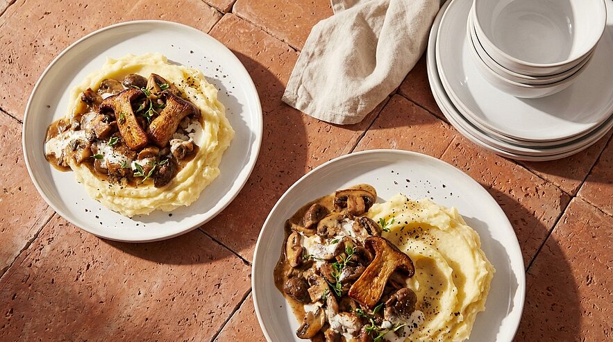 Zwei Teller mit cremigem Kartoffelpüree, darauf ein herzhaftes Pilzragout mit Kräutern und frisch gemahlenem schwarzem Pfeffer.