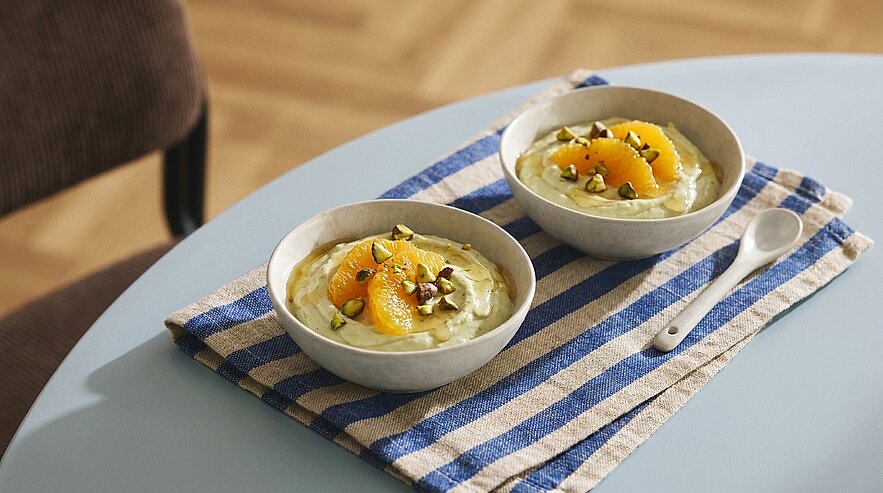 Zwei Schalen mit cremigem Porridge, jeweils garniert mit Orangenfilets und Kürbiskernen, auf einem blau-weiß gestreiften Tuch auf einem Tisch.