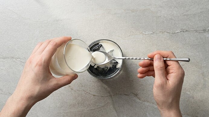Zwei Hände gießen Milch in ein Glas mit Eiswürfeln und dunklem Kaffee und rühren den Inhalt mit einem langen Löffel.