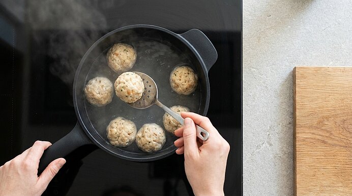Draufsicht: Hände, die runde Knödel in einem Topf mit kochendem Wasser garen; einer wird mit einem Schaumlöffel herausgehoben.