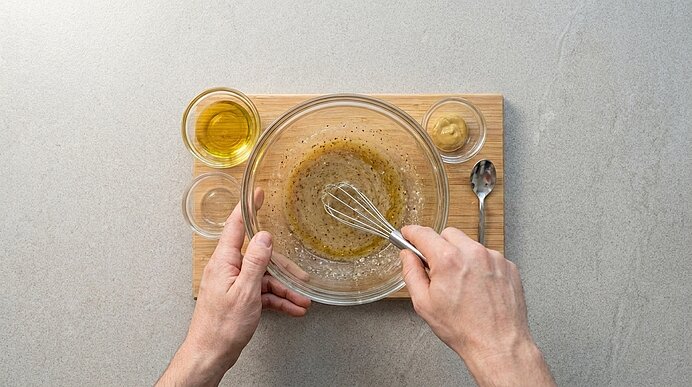 Zwei Hände rühren mit einem Schneebesen in einer Glasschüssel eine Vinaigrette; auf einem Holzbrett daneben kleine Schälchen mit Öl, Essig und Senf sowie ein Löffel.