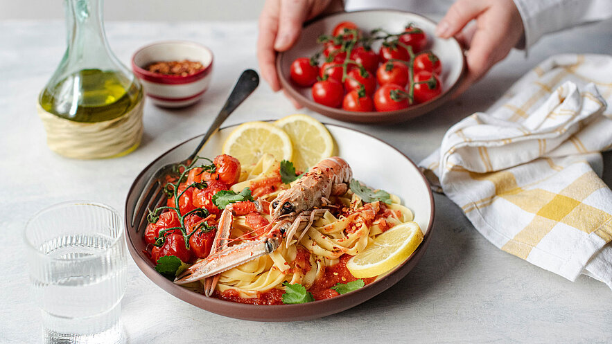Ein Teller Pasta mit einer Tomatensauce, darauf große Garnelen und frische Kirschtomaten. Garniert mit Zitronenscheiben und frischem Koriander. Eine Hand hält eine zusätzliche Schale mit Kirschtomaten, während ein Glas Wasser und eine Flasche Olivenöl im Hintergrund sichtbar sind.
