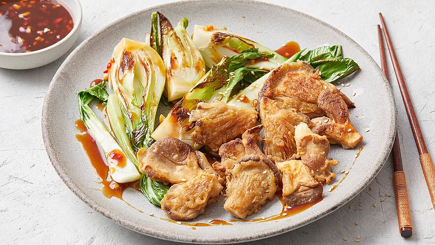 Ein Teller mit gebratenem Fleischstücken und sautiertem Pak Choi, serviert mit einer dunklen Soße. Neben dem Teller befinden sich Essstäbchen.