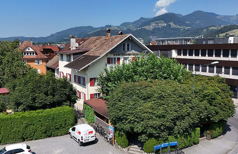 Das Bild zeigt eine städtische Umgebung mit verschiedenen Gebäuden. Ein helles zweigeschossiges Haus mit roten Fenstern und einem angrenzenden Garten ist prominent. Im Hintergrund erheben sich grüne Berge unter einem klaren blauen Himmel. Ein Fahrzeug steht auf einem gepflasterten Parkplatz.
