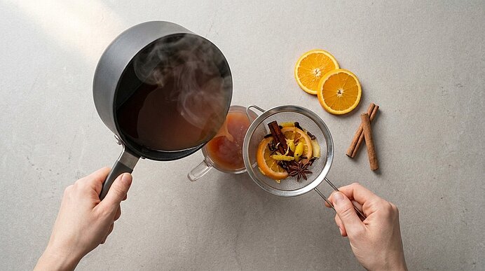 Zwei Hände sieben dampfenden Glühwein aus einem Topf in einen Glasbecher; im Sieb Orangen- und Zitronenscheiben mit Gewürzen (Zimtstange, Sternanis). Daneben liegen auf der Arbeitsfläche Orangenscheiben und zwei Zimtstangen.