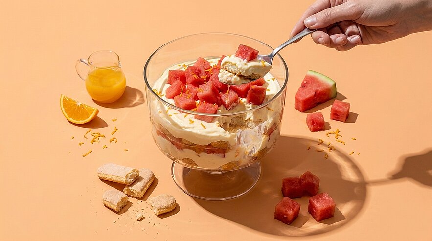 Glasschale mit geschichtetem Dessert aus Creme und Biskuit, oben mit frischen Wassermelonenwürfeln; Hand führt einen Löffel zum Servieren.