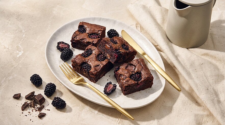 Drei saftige Schokoladenbrownies mit eingebackenen und dekorativen Brombeeren auf einem weißen Teller, daneben goldene Gabel und Messer sowie einige frische Brombeeren und Stücke dunkler Schokolade.