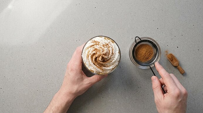 Eine Hand hält ein Glas Iced Spiced Latte mit Sahne und Zimt, die andere hält ein kleines Sieb mit Zimtpulver.