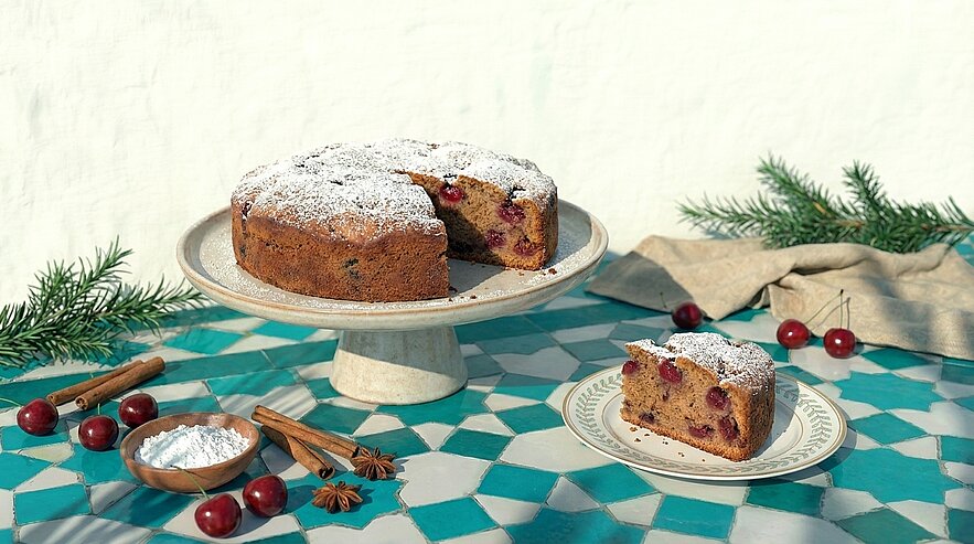 Ein frisch gebackener Kuchen mit Kirschen, bestäubt mit Puderzucker, steht auf einem eleganten Teller. Ein Stück des Kuchens ist auf einem Unterteller daneben platziert. Die Szene wird durch einige Zimtstangen, Puderzucker und grüne Zweige ergänzt. Der Tisch hat ein geometrisches Muster.