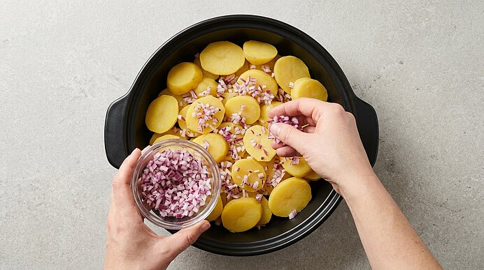 Zwei Hände streuen fein gewürfelte rote Zwiebel über in Scheiben geschnittene Kartoffeln in einem schwarzen Topf.