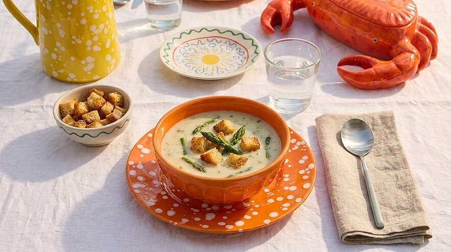 Cremige Spargelsuppe in orangefarbener Schale, garniert mit Spargelspitzen und Croutons, auf gedecktem Tisch mit Glas Wasser, Löffel und Serviette.