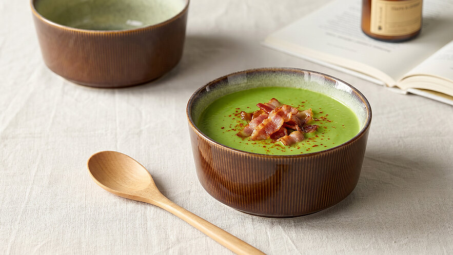 Eine grüne Suppe in einer braunen Schüssel, garniert mit knusprigem Speck und roten Gewürzen. Neben der Schüssel liegt ein Holzlöffel. Im Hintergrund sind weitere Schüsseln und ein aufgeschlagenes Buch zu sehen.