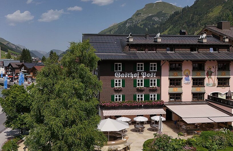 Ein traditioneller Gasthof namens "Gasthof Post" in einer malerischen Berglandschaft. Das Gebäude ist liebevoll gestaltet mit bunt blühenden Fenstern und Wandmalereien. Vor dem Gasthof befinden sich Terrassen mit Sonnenschirmen und Tischen, umgeben von Bäumen und einer schönen Umgebung.