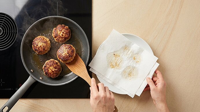 Vier gebratene Frikadellen in einer Pfanne; eine Hand hebt mit Holzspatel eine Frikadelle, die andere hält einen mit Küchenpapier ausgelegten Teller zum Abtropfen des Fettes.