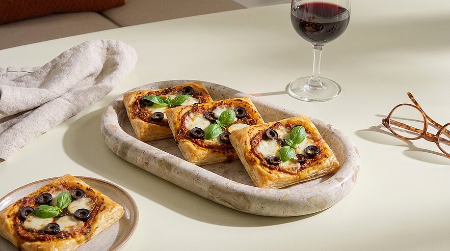 Drei quadratische Mini‑Pizzen mit geschmolzenem Käse, schwarzen Olivenscheiben und frischen Basilikumblättern auf einer ovalen Marmorplatte, daneben eine weitere Mini‑Pizza auf einem kleinen Teller.