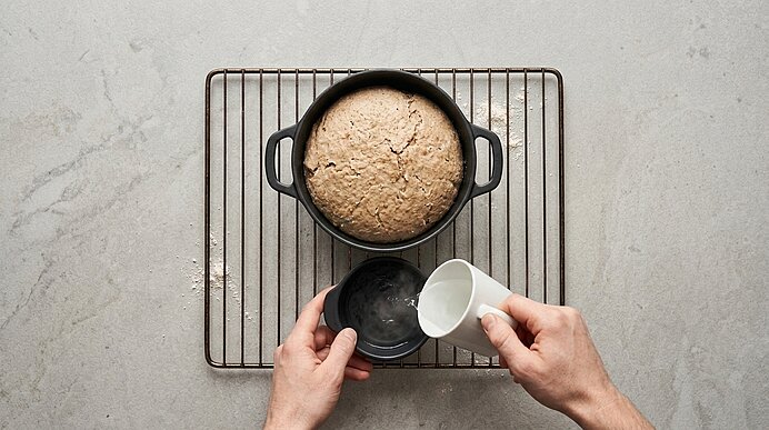 Zwei Hände gießen Wasser aus einer Tasse in eine kleine Schale neben der Backform mit dem Roggenbrot.