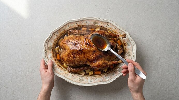 Zwei Hände halten eine ovale Servierplatte mit einem goldbraun gebratenen Brathähnchen auf geröstetem Gemüse; eine weitere Hand löffelt Bratensaft über das Geflügel.