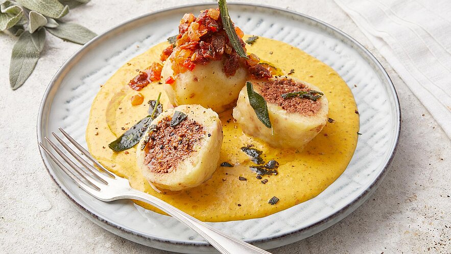 Ein Teller mit gefüllten Kartoffelknödeln, die in einer goldgelben Sauce liegen. Die Knödel sind mit einer roten, würzigen Mischung garniert und mit Salbeiblättern dekoriert. Eine Gabel liegt neben dem Teller.