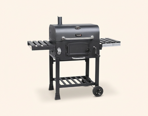 Ein schwarzer Grill mit zwei seitlichen Ablageflächen und einem Untergestell auf Rollen. Der Grill hat eine geschlossene Kammer mit einer Tür und einem Kamin auf der Oberseite. Die Konstruktion ist stabil und eignet sich für das Grillen im Freien.