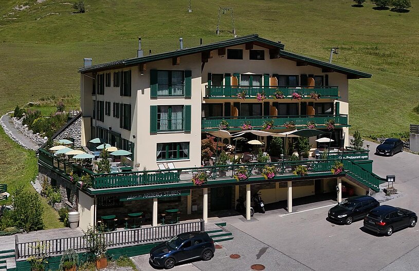 Ein mehrstöckiges Gebäude mit grünem Balkon und blühenden Pflanzen. Vor dem Gebäude stehen Tische mit Sonnenschirmen. Parkplätze sind im Vordergrund sichtbar. Die Umgebung besteht aus einer grünen Wiese mit sanften Hügeln im Hintergrund.