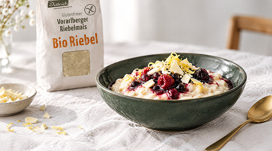 Grüne Schüssel mit cremigem Maisgrieß-Riebel, garniert mit Beerenkompott und Zitronenzesten; daneben Packung Bio Riebel (glutenfreier Vorarlberger Riebelmais).
