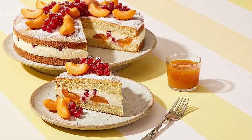 Gestapelte Biskuit-Cremetorte mit Fruchtfüllung, garniert mit Aprikosenhälften und roten Johannisbeeren; ein Stück auf einem Teller, daneben ein Glas Aprikosenmarmelade und eine Gabel.