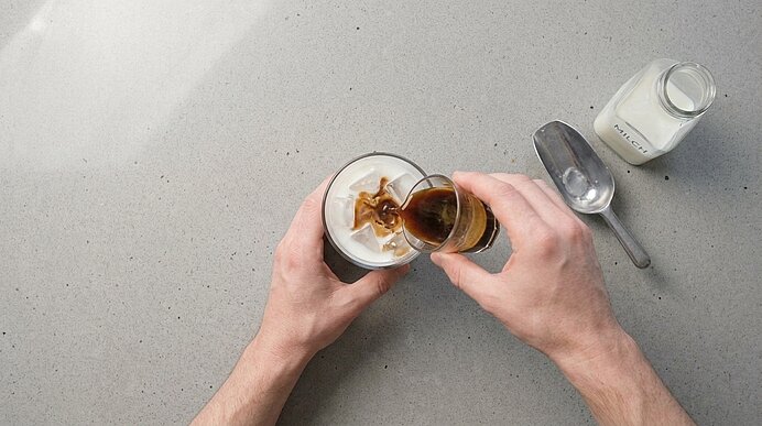 Zwei Hände gießen Espresso aus einem kleinen Glas in ein Glas mit kalter Milch und Eiswürfeln.