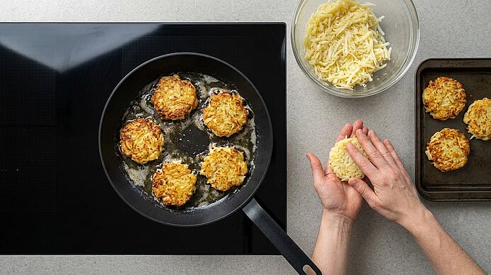 Zwei Hände formen Kartoffelrösti, während weitere Rösti in einer Pfanne auf dem Kochfeld goldbraun braten.