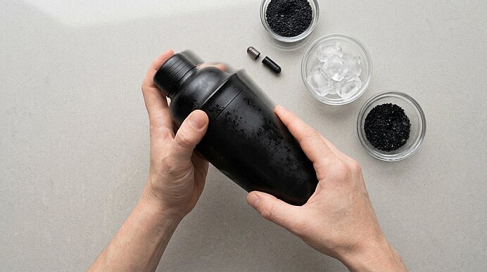 Zwei Hände schütteln einen mattschwarzen Cocktailshaker. Daneben stehen kleine Schalen mit Eis und schwarzem Salz.