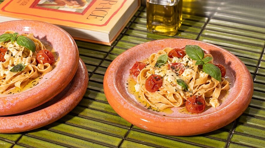 Zwei Teller mit frischen Tagliatelle, garniert mit Kirschtomaten und frischem Basilikum, stehen auf einem grünen Tisch. Im Hintergrund liegt ein Kochbuch und eine Flasche Olivenöl. Die Pastagerichte wirken appetitlich und einladend.