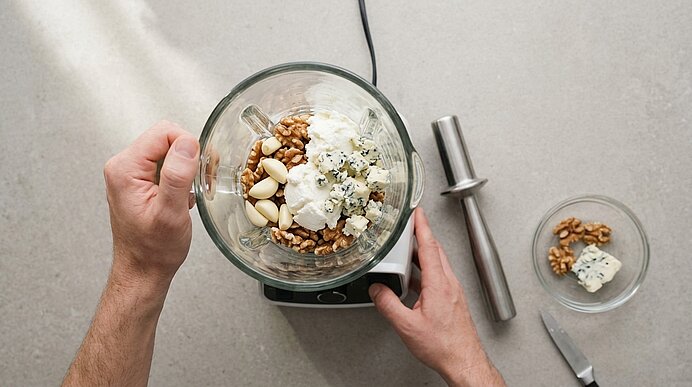Zwei Hände bedienen einen Standmixer, in dem sich Walnüsse, Knoblauch, Ricotta und Gorgonzola befinden.