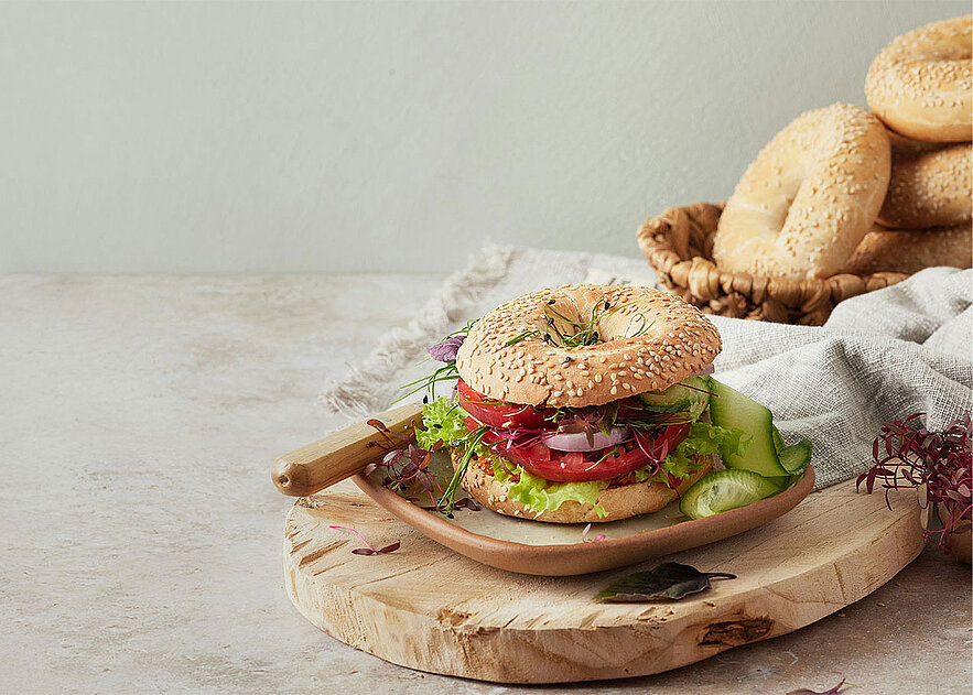 Ein Sesam-Bagel-Sandwich mit Salat, Tomaten, Gurken, Zwiebeln und Sprossen auf einem Holzteller. Im Hintergrund steht ein Korb mit einfachen Bagels.
