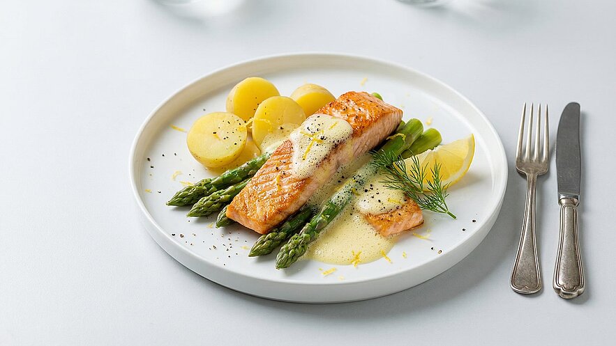Ein Teller mit gebratenem Lachs, serviert mit grünem Spargel, Kartöffelchen und einer Zitronensauce. Das Gericht ist mit frischem Dill und Zitronenscheiben garniert. Rechts liegen eine Gabel und ein Messer.