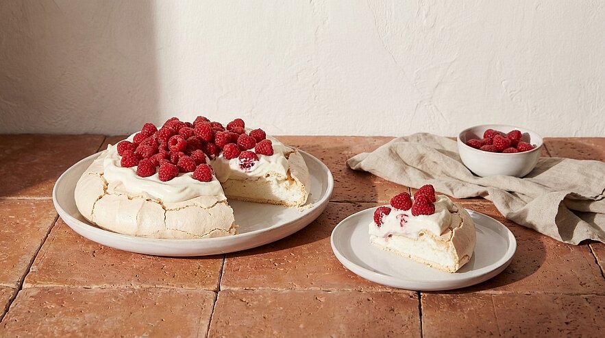 Baiser-Torte mit knusprigem Baiserboden, cremiger Schlagsahne und frischen Himbeeren auf einem Teller; ein Stück auf einem kleinen Teller daneben, Schale mit zusätzlichen Himbeeren.