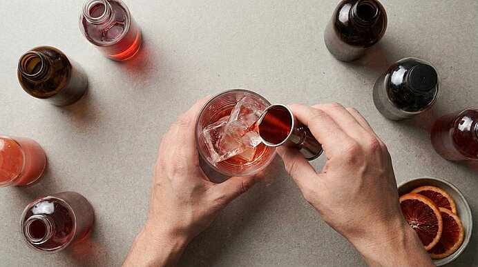 Zwei Hände gießen mit einem Messbecher ein rotes Getränk über Eis in ein Cocktailglas; rundherum stehen mehrere kleine Flaschen und Blutorangenscheiben.