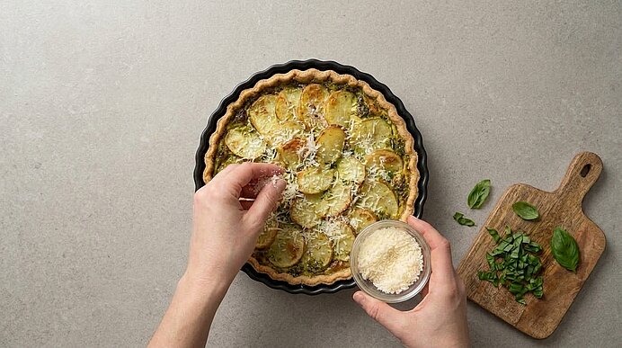 Zwei Hände bestreuen eine gebackene Kartoffeltarte mit geriebenem Parmesan aus einer kleinen Glasschale.