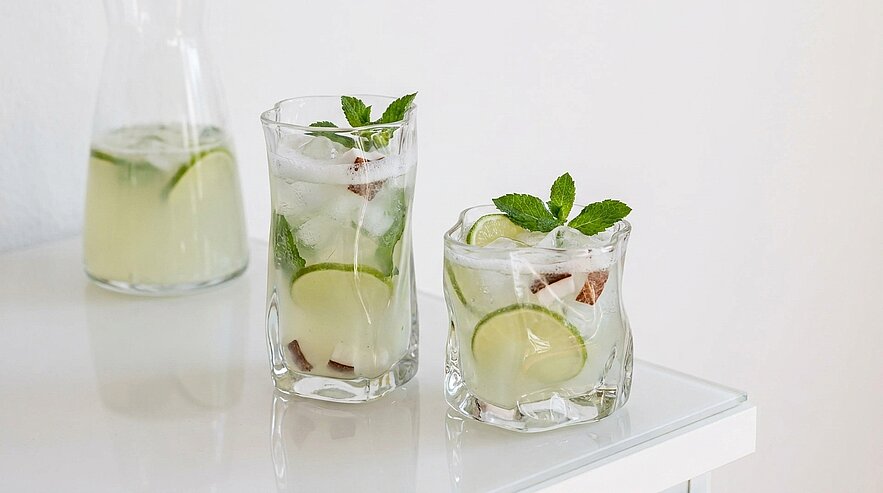 Zwei Gläser mit eisgefülltem Limetten‑Minz‑Getränk (Limettenscheiben, Minzblätter, Eiswürfel) auf weißer Oberfläche; im Hintergrund eine Karaffe mit dem gleichen Getränk.