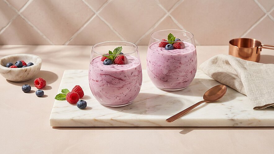 Zwei Gläser mit einem rosa Smoothie, garniert mit frischen Himbeeren und Heidelbeeren sowie einem Minzblatt. Sie stehen auf einem Marmortablett, begleitet von einer kleinen Schüssel mit Beeren und einem Löffel. Hintergrund in sanften, neutralen Farben.
