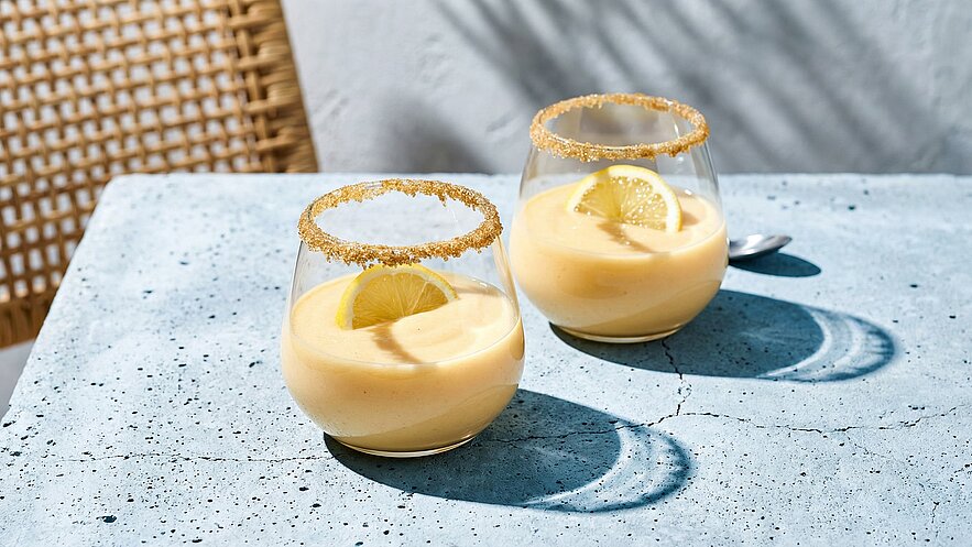 Zwei Gläser mit einem cremigen, gelben Dessert stehen auf einem hellen Tisch. Jedes Glas hat einen Rand aus braunem Zucker und ist mit einer Zitronenscheibe garniert. Die Szenerie vermittelt eine frische, einladende Atmosphäre.