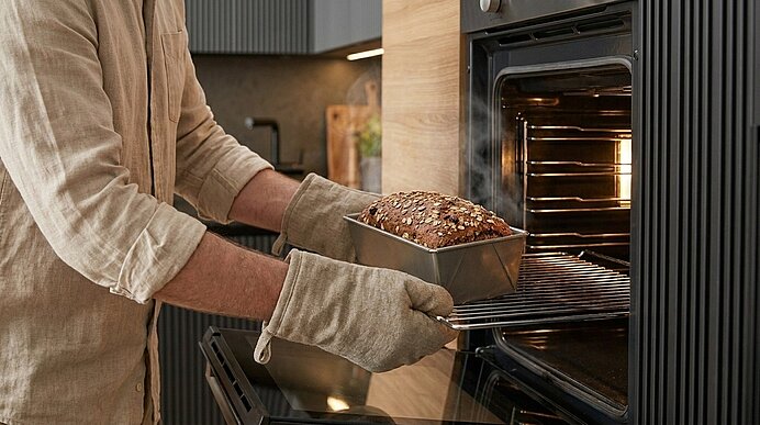 Person mit Ofenhandschuhen entnimmt ein frisch gebackenes Mehrkorn-Kastenbrot in einer Metallform aus einem Backofen.