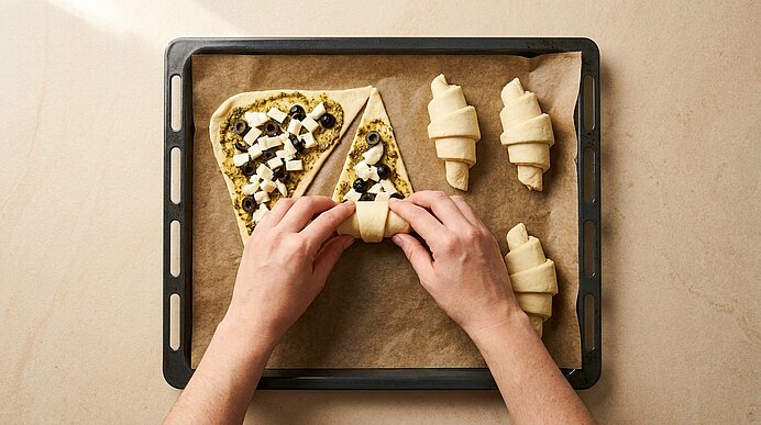 Zwei Hände rollen auf einem mit Backpapier belegten Backblech belegte Teigdreiecke (Pesto, Oliven, Feta) zu Croissant‑Hörnchen; drei fertige Hörnchen liegen daneben.