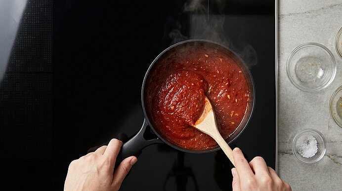 Zwei Hände rühren mit einem Holzlöffel dickflüssige Tomatensauce in einem schwarzen Stieltopf auf dem Kochfeld.