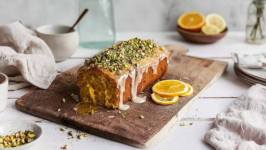 Ein saftiger, mit Zuckerguss überzogener Zitronenkuchen liegt auf einem Holzbrett. Die Oberfläche ist mit gehackten Pistazien dekoriert. Neben dem Kuchen befinden sich frische Zitronenscheiben. Der Hintergrund ist hell und schlicht, wodurch der Kuchen im Vordergrund steht.