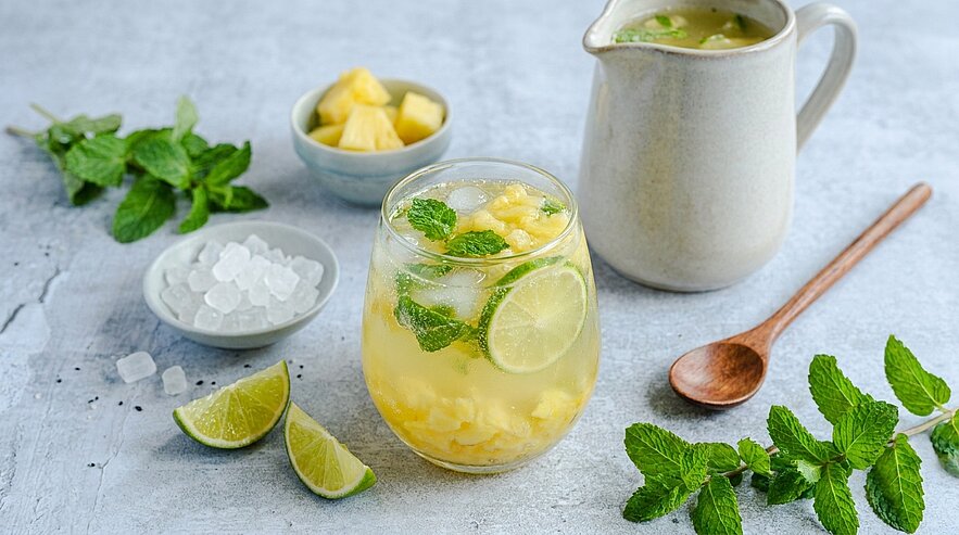 Ein Glas erfrischendes Getränk mit Ananasstücken, Limettenscheiben und frischen Minzblättern, umgeben von Eiswürfeln und einer kleinen Kanne mit einem weiteren Getränk. Im Hintergrund sieht man einen kleinen Schalen mit Ananasstücken und frischen Minzblättern.