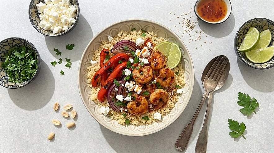 Schüssel mit gewürzten Garnelen auf Couscous, dazu geröstete rote Paprika und Zwiebelringe, zerbröselter Feta, Limettenscheibe und frische Kräuter, Besteck daneben.