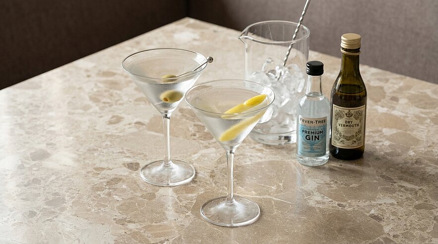 Zwei klassische Martinigläser mit klaren Cocktails, eines garniert mit einer Olive, das andere mit einer Zitronenzeste; daneben kleine Gin- und Wermutflaschen sowie ein Eisbehälter mit Barlöffel auf einer Marmoroberfläche.